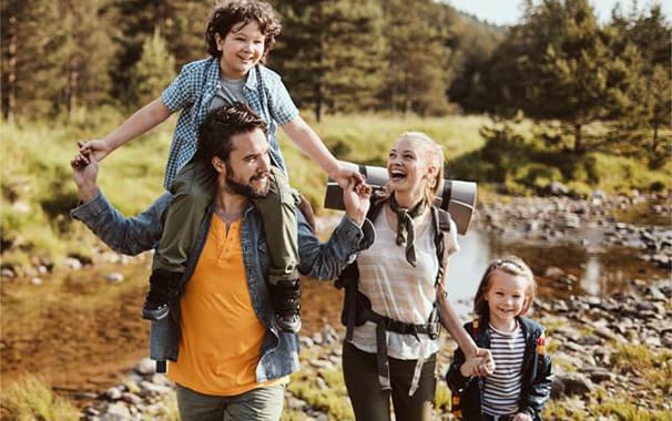 A family of four is walking outside. The father has one child riding on his shoulders, the other child is next to the mom, holding her hand. The family looks to be happy and having fun