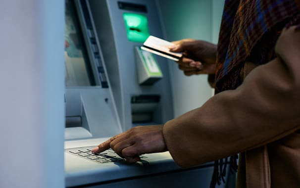 Photo of a person standing at an ATM about to insert their card. The photo is focused on the person's hands holding the debit card.