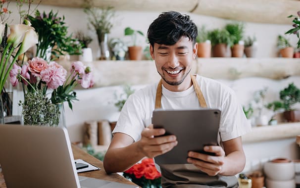 A man is at work, and is wearing an apron, he is holding a tablet and is looking at its screen and smiling