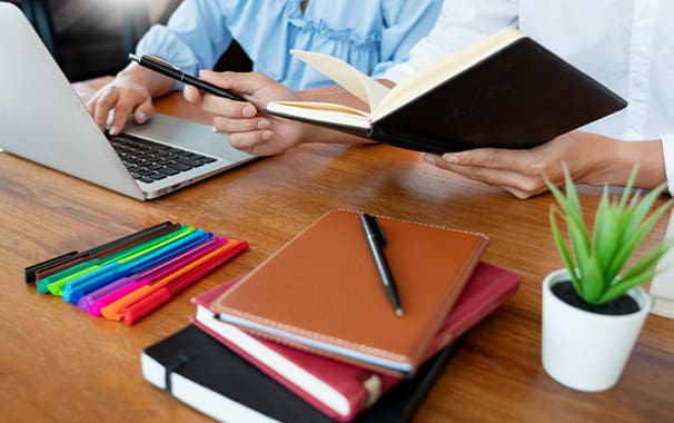 Photo of a desk. On the desk is a stack of notebooks with a pen on top. In the background, you see a person working on their laptop.