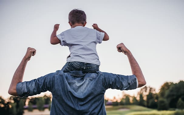 Photo taken from behind and a father has his young soon sitting on his shoulders. Both of them have their arms flexed in a body builder pose