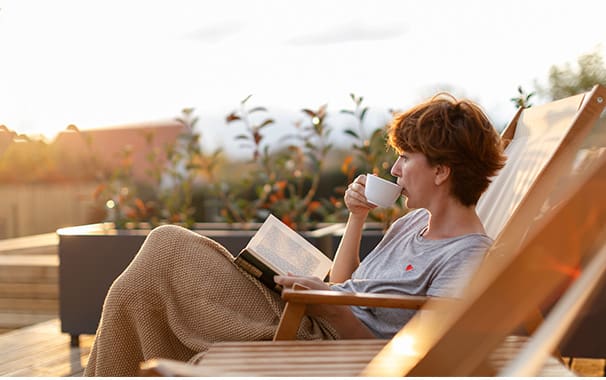 A woman is seated outside in a lounge chair, reading a book and sipping a cup of tea