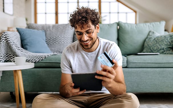 A man is seated on the floor in front of his sofa and he is holding a tablet in his hands