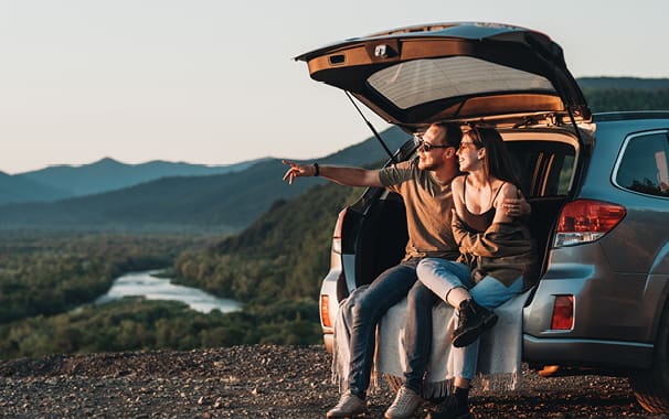A man and woman sit on the back of their car with the trunk open, staring at a scenic view